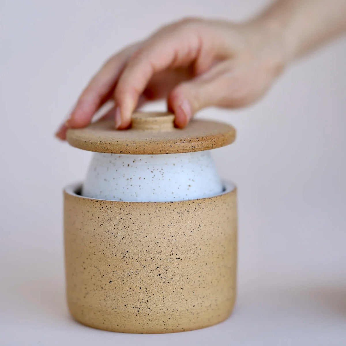 Hand closing a ceramic jar with a wooden lid on a light background
