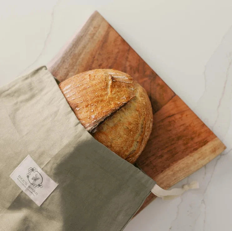 A light olive green linen bread bag with a white brand label, partially holding a round rustic loaf of bread on a wooden board over a white marble surface.