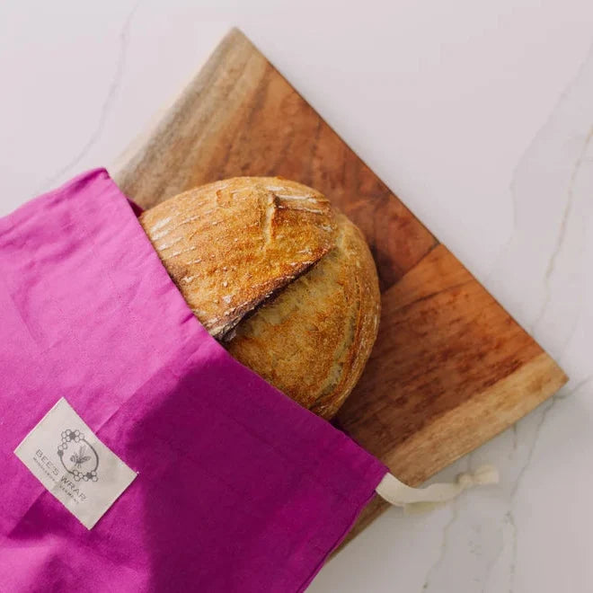 A round artisan bread loaf partially tucked into a bright magenta reusable fabric bag with a white brand label, placed on a wooden board over a light marble surface.