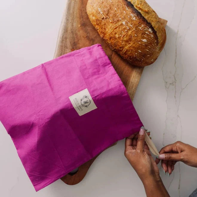 Person opening a pink fabric package on a wooden board with bread in the background.
