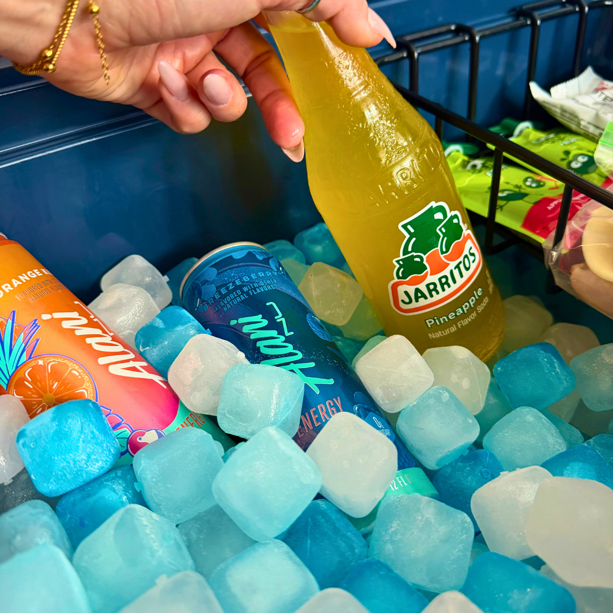 Person holding a Jarritos pineapple soda bottle over ice cubes with other drinks in the background.