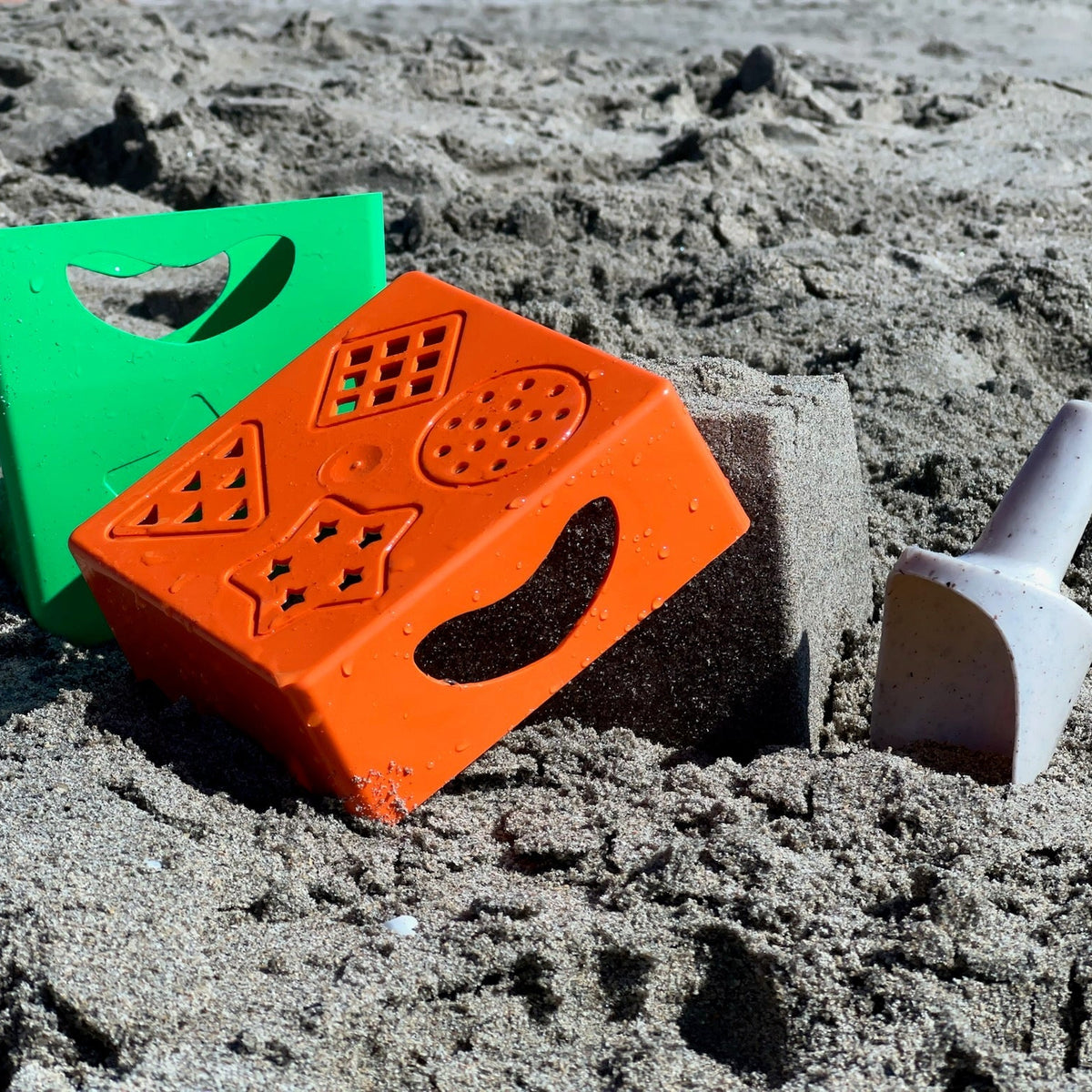A set of plastic sand toys on a sandy beach, including an orange mold, a green scoop, and a gray shovel.