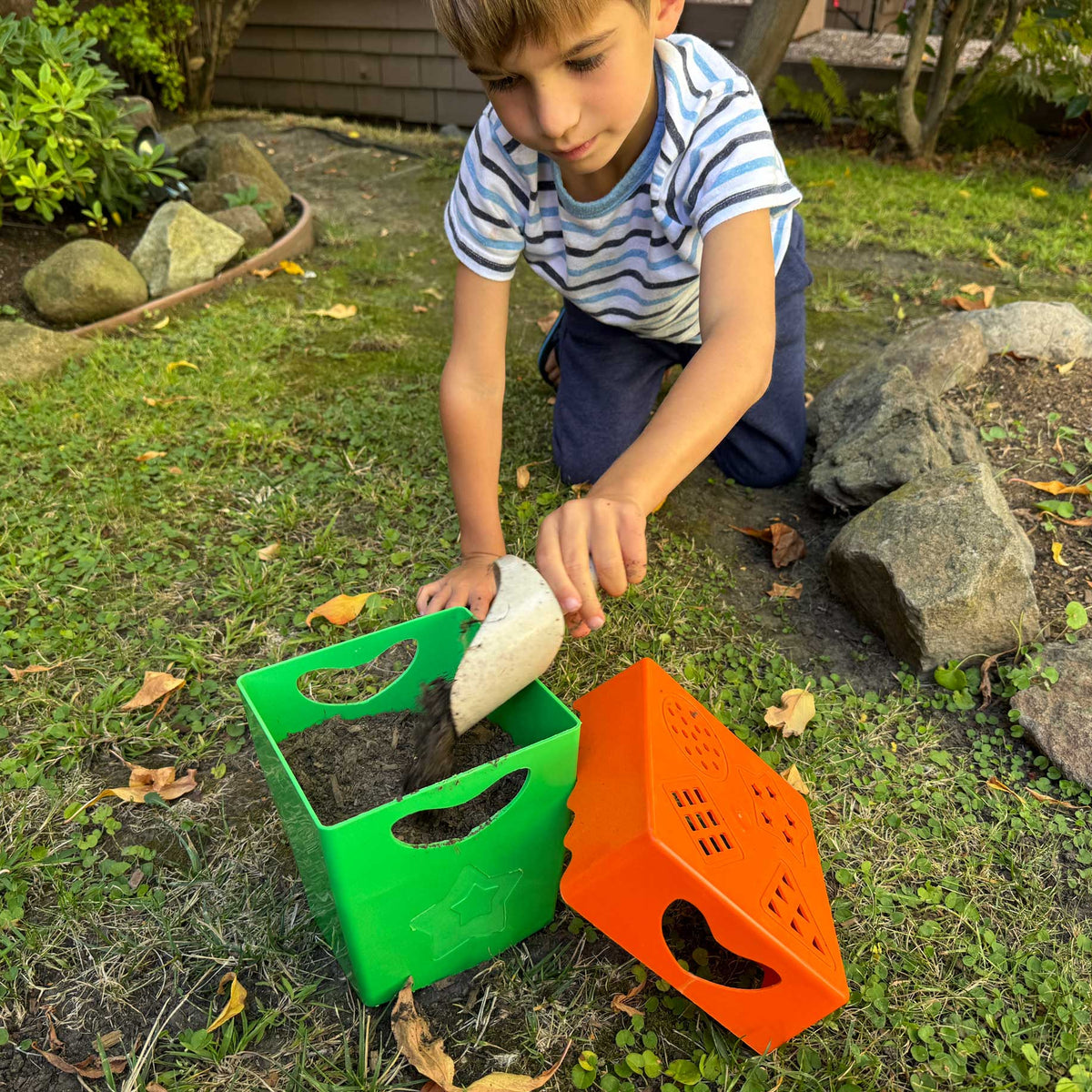 A young boy kneeling in a grassy backyard garden, using a small white scoop to fill a green plastic planter box. An orange plastic planter box lies nearby on the grass.