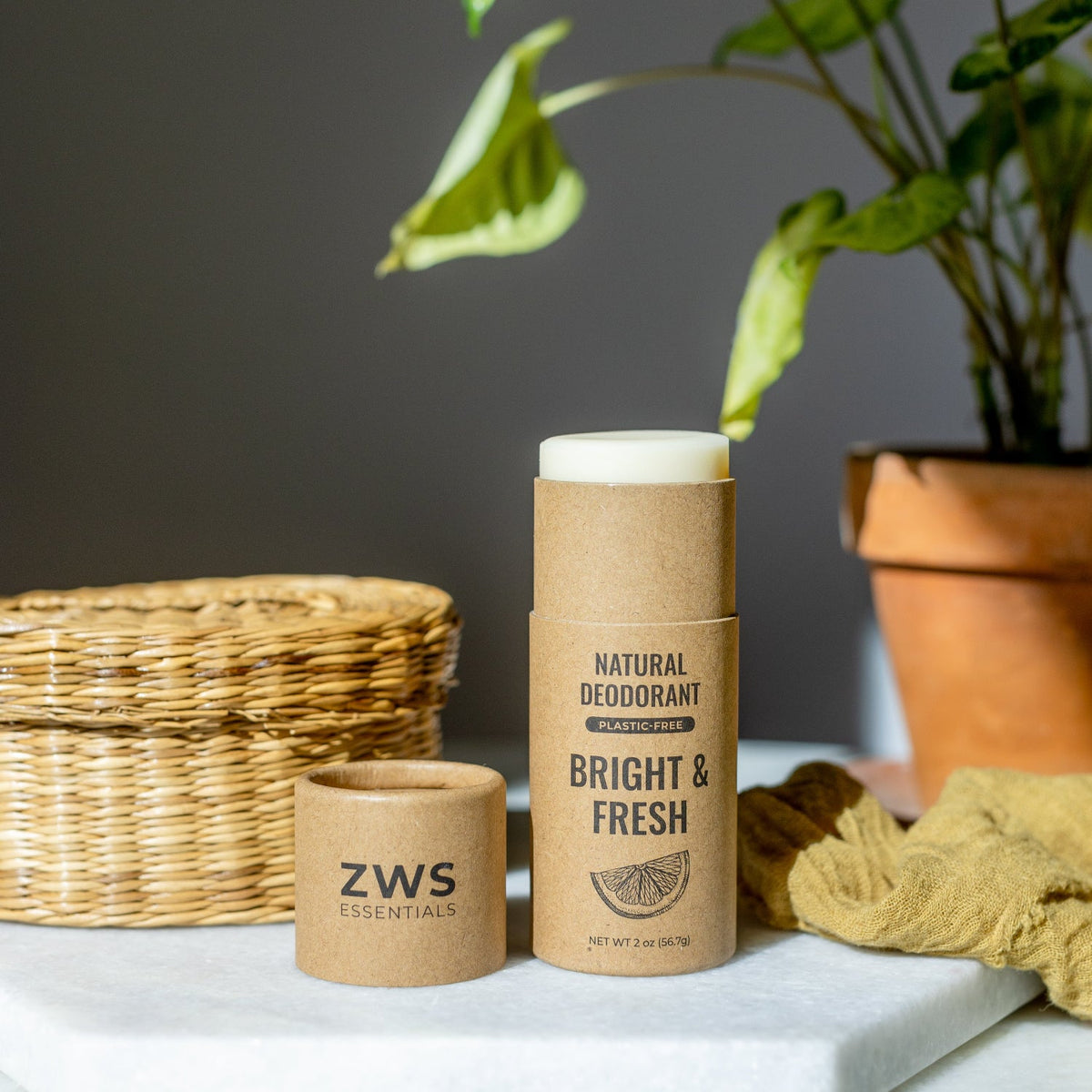 A natural deodorant stick in a kraft paper sleeve stands on a white marble surface next to its open cap, with a wicker basket, potted plant, and mustard cloth in the background.