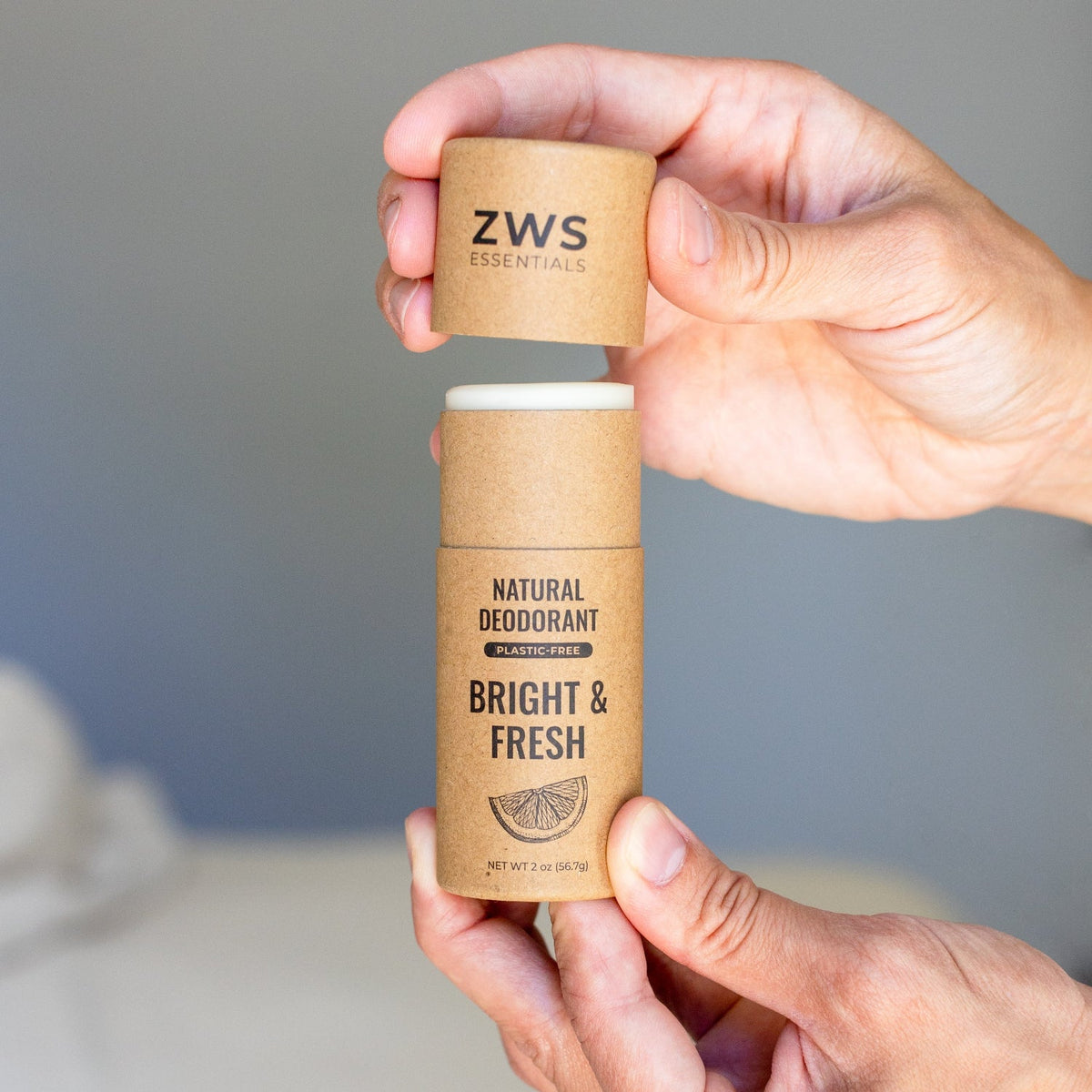 A person's hands holding a ZWS Essentials natural deodorant stick in a brown kraft paper tube, with the cap removed and held above the container. The background is a soft, out-of-focus gray and white interior.
