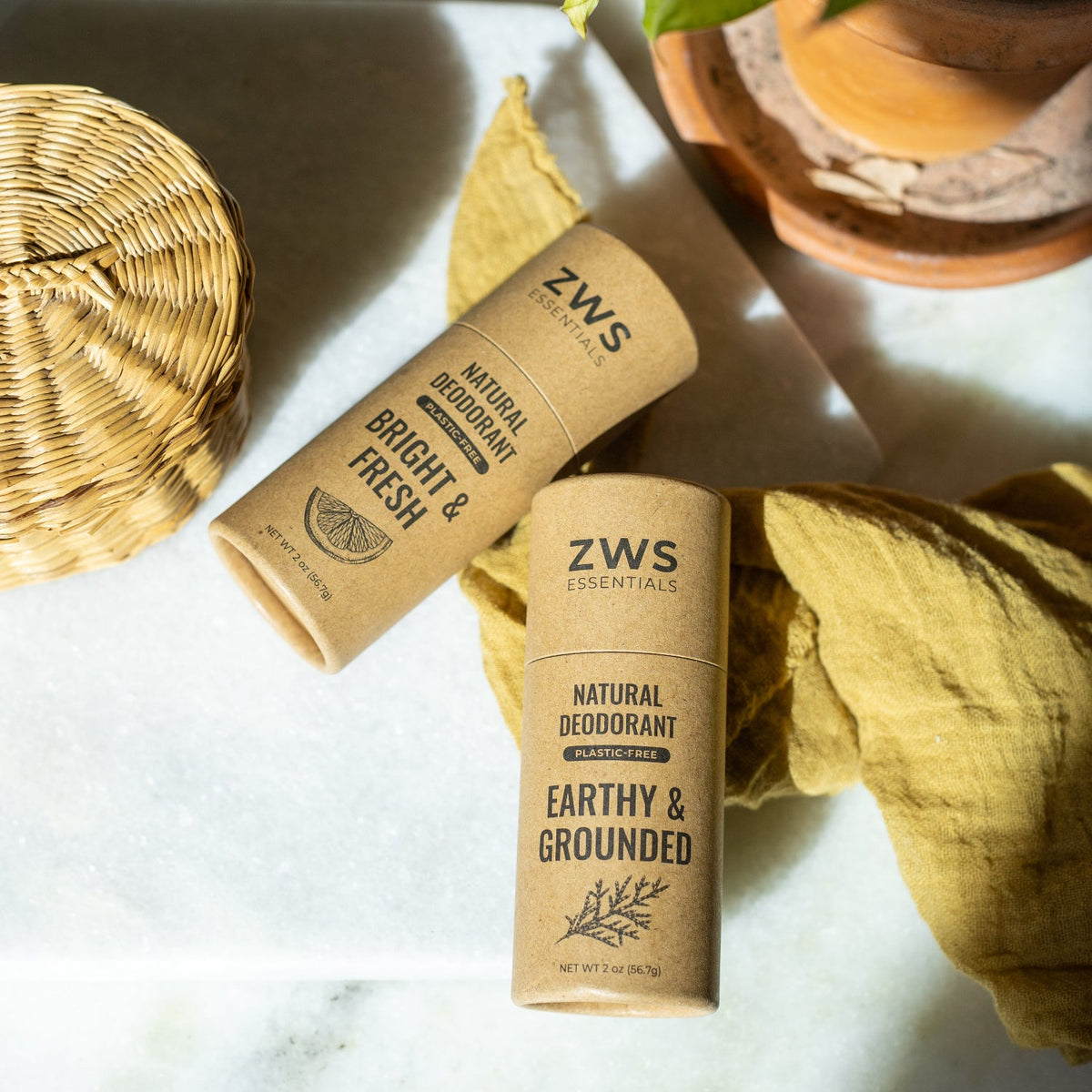 Two ZWS Essentials natural deodorant sticks in kraft paper packaging, labeled 'Bright & Fresh' and 'Earthy & Grounded,' arranged on a light marble surface with a yellow cloth, woven basket, and terracotta pot in the background.