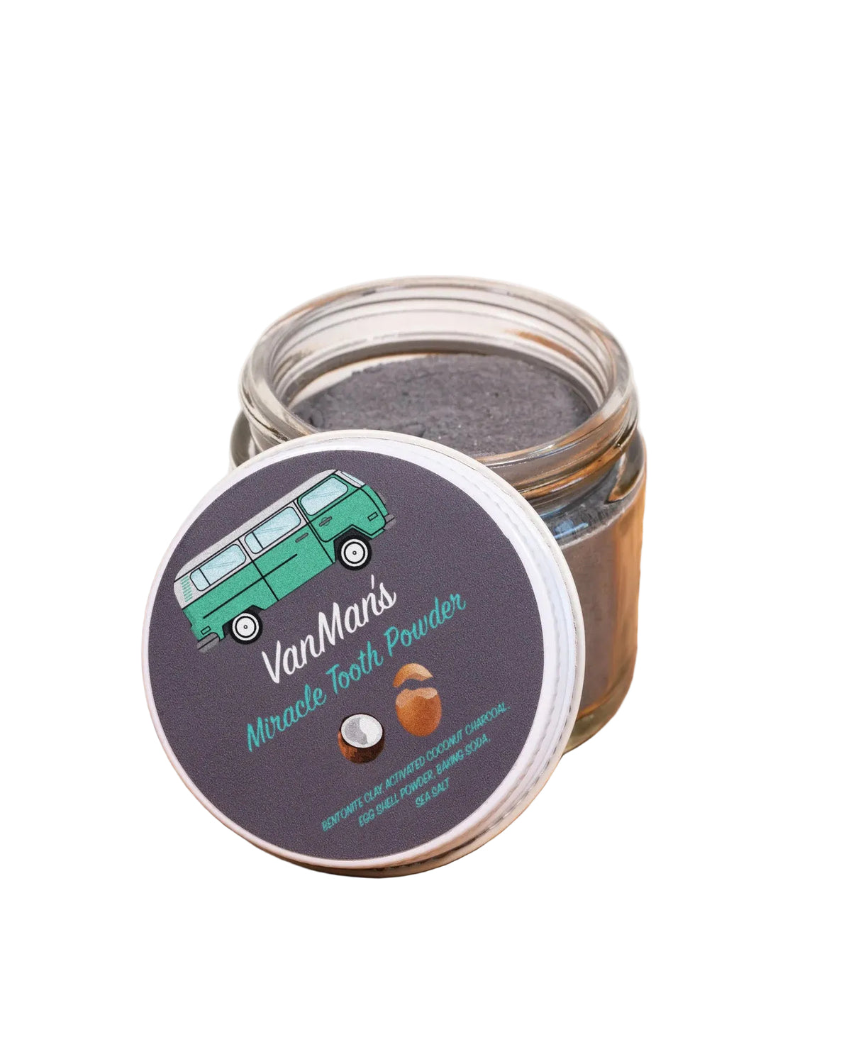 A small clear glass jar filled with dark gray tooth powder sits on a wooden surface, with the round lid placed in front. The lid has a dark label reading "VanMan's Miracle Tooth Powder" with a green van illustration and a small orange-and-white coconut graphic.