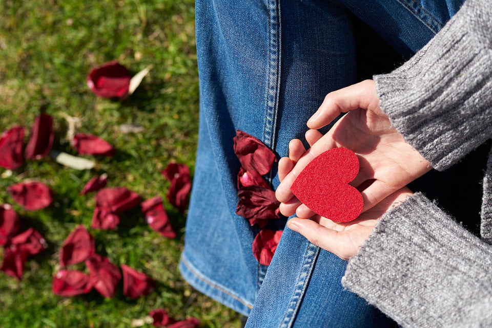 Person holding a red heart-shaped object with fallen red petals on grass