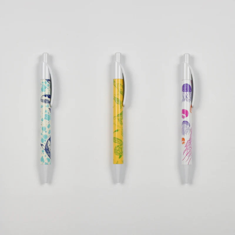 Three retractable ballpoint pens with clear plastic tips and clips, featuring colorful printed patterns in blue, green, yellow, purple, and pink on a light gray background.
