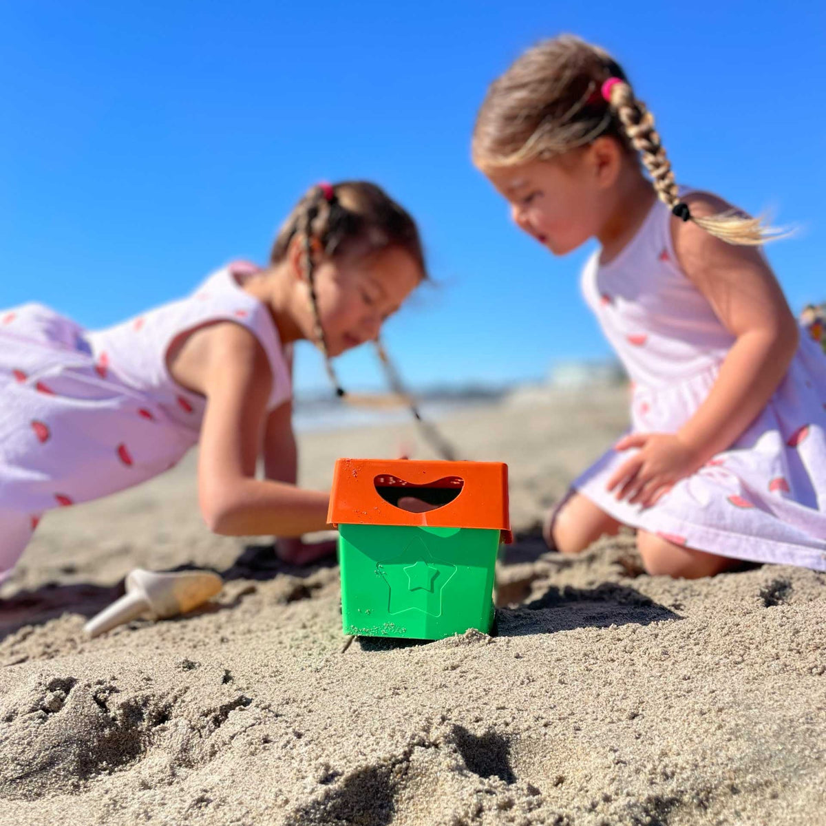 Two young girls in white dresses with pink watermelon prints are playing in the sand on a beach, looking at a small green and orange toy box.