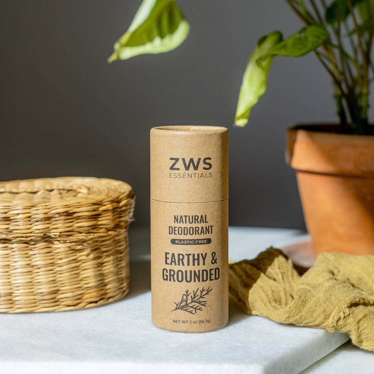 ZWS Essentials Earthy & Grounded Natural Deodorant in a kraft paper cylindrical tube, styled on a marble surface with a wicker basket, yellow cloth, and potted plant in the background.