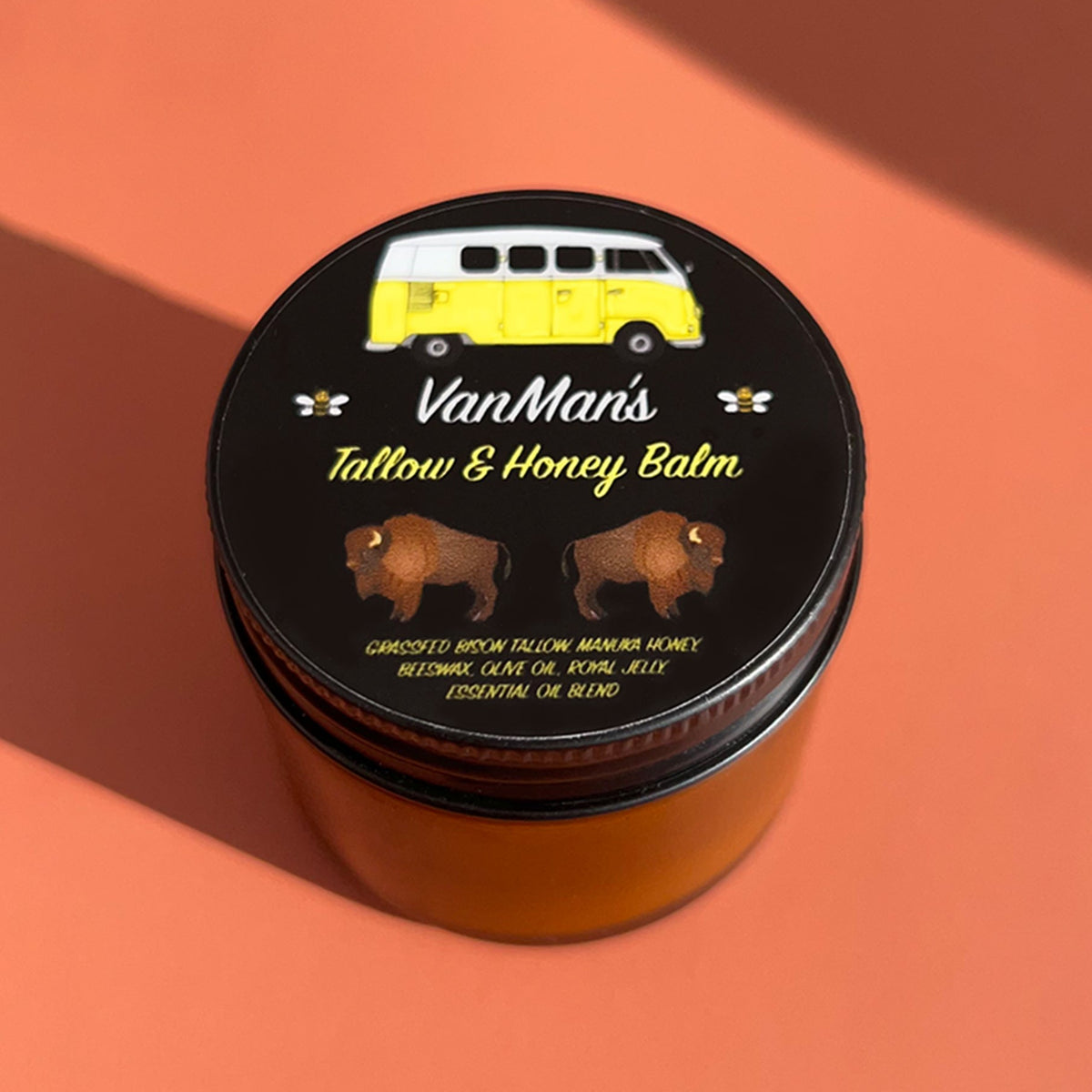 A round tin of Van Man's Tallow & Honey Balm with a black lid featuring a yellow van illustration and bison graphics, set against a warm peach background with diagonal shadows.